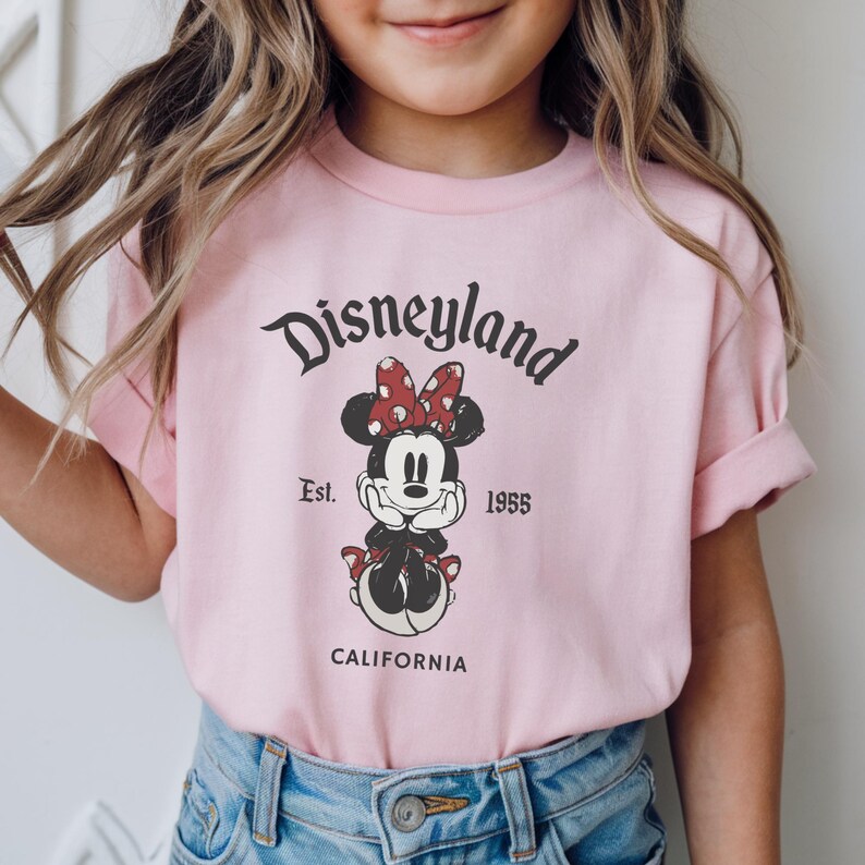 May include: Pink t-shirt featuring a Minnie Mouse graphic with a red bow and the text "Disneyland Est. 1955 CALIFORNIA". The shirt is made of a soft material and is suitable for children.