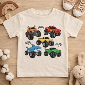 May include: Cream-coloured short-sleeved t-shirt with a graphic of five monster trucks. The trucks are orange, red, yellow, blue, and green, each with large black tyres and checkered flags. A fun design for children.