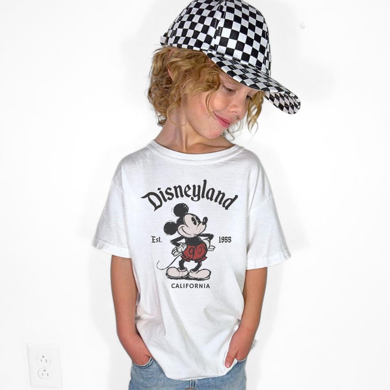 May include: A white t-shirt with a Disneyland graphic featuring Mickey Mouse, paired with a black and white checkered baseball cap. The shirt has the text "Disneyland Est. 1955 California".