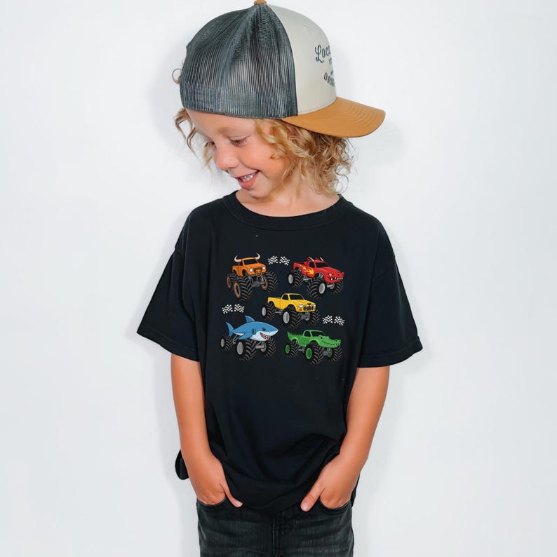 May include: A black t-shirt with colourful monster truck graphics. The shirt shows six different monster truck designs, including a shark-themed truck. The child is also wearing a trucker hat with a grey mesh back and a brown brim.