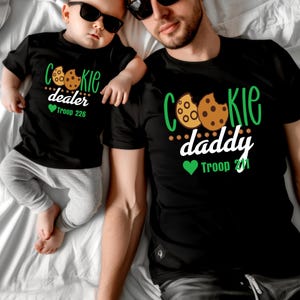Peut inclure: Deux t-shirts noirs avec des graphiques sur le th&egrave;me des cookies. L'un dit "Cookie dealer" et "Troop 226". L'autre dit "Cookie daddy" et "Troop 221". Les deux chemises ont un motif de cookie.