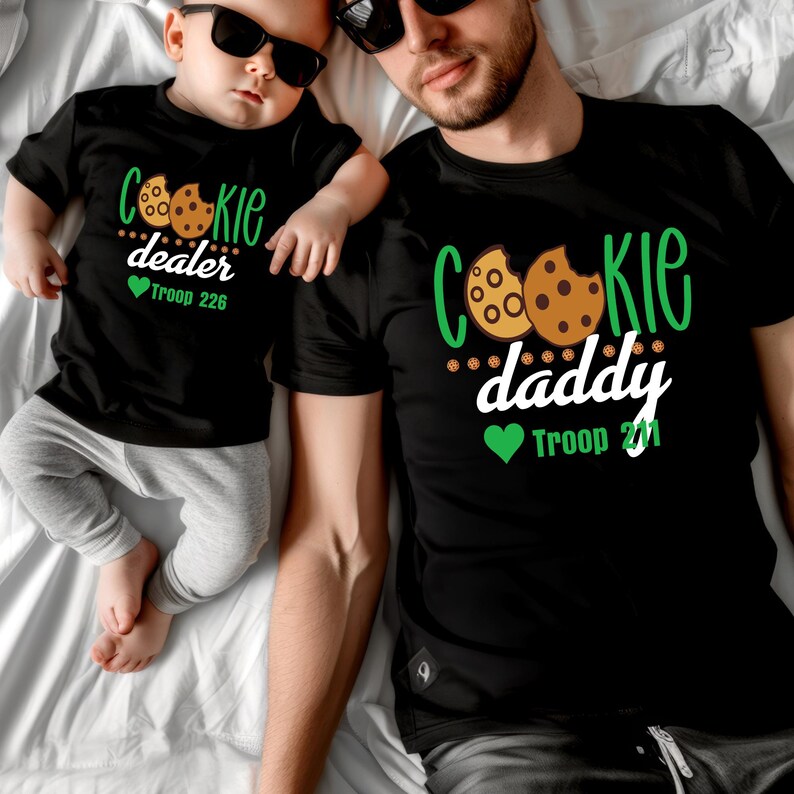 Peut inclure: Deux t-shirts noirs avec des graphiques sur le th&egrave;me des cookies. L'un dit "Cookie dealer" et "Troop 226". L'autre dit "Cookie daddy" et "Troop 221". Les deux chemises ont un motif de cookie.