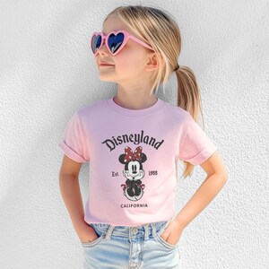 May include: A young person wearing a pink t-shirt with the Disneyland logo and a Minnie Mouse graphic. The shirt also has the text "Est. 1955" and "California". The person is wearing pink heart-shaped sunglasses and blue jeans.