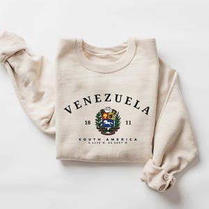 May include: A cream-coloured sweatshirt featuring the word "VENEZUELA" in black, arched across the top. Below is a colourful emblem, the words "SOUTH AMERICA" and coordinates. The sleeves are rolled up.