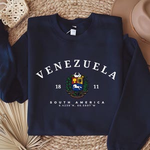 May include: A navy blue sweatshirt featuring the word "VENEZUELA" in white arched lettering. Below is a coat of arms, the words "SOUTH AMERICA" and coordinates. The sweatshirt is displayed on a woven surface.