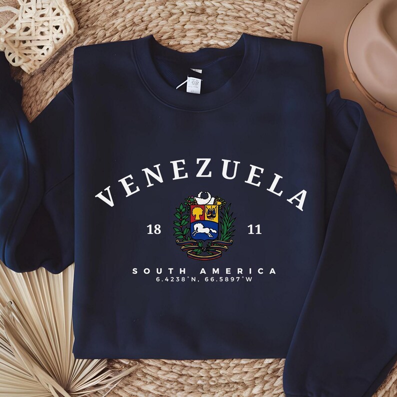 May include: A navy blue sweatshirt featuring the word "VENEZUELA" in white arched lettering. Below is a coat of arms, the words "SOUTH AMERICA" and coordinates. The sweatshirt is displayed on a woven surface.