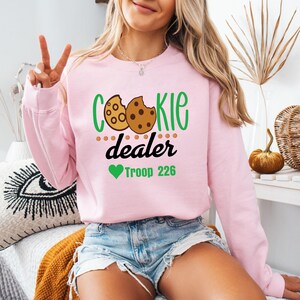 Peut inclure: Un sweat-shirt rose clair avec les mots "Cookie Dealer" en vert et noir, ainsi qu'un dessin de biscuit. En dessous du texte, un c&oelig;ur vert et les mots "Troop 226".