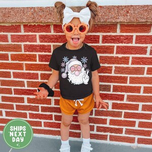 May include: A child in a black Santa Claus graphic t-shirt, orange shorts, and orange sunglasses. The Santa graphic shows him blowing a bubble and giving a peace sign. The child is standing in front of a brick wall.