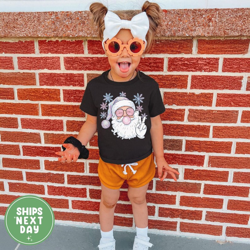 May include: A child in a black Santa Claus graphic t-shirt, orange shorts, and orange sunglasses. The Santa graphic shows him blowing a bubble and giving a peace sign. The child is standing in front of a brick wall.