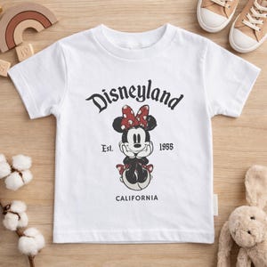 May include: White t-shirt featuring a Minnie Mouse graphic with a red polka dot bow and the text "Disneyland" above the character. The shirt also includes the text "Est. 1955" and "CALIFORNIA".