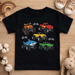 May include: Black t-shirt with illustrations of monster trucks in various colours. The trucks are orange, red, yellow, blue, and green, each with animal-themed designs. Checkered flags are positioned near each truck, implying a racing theme.