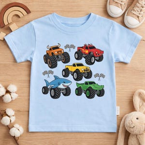 May include: A pale blue t-shirt displaying five monster trucks in orange, red, yellow, blue, and green. Each truck has large black tyres and a unique design, including a shark and an alligator. Checkered flags are also included.