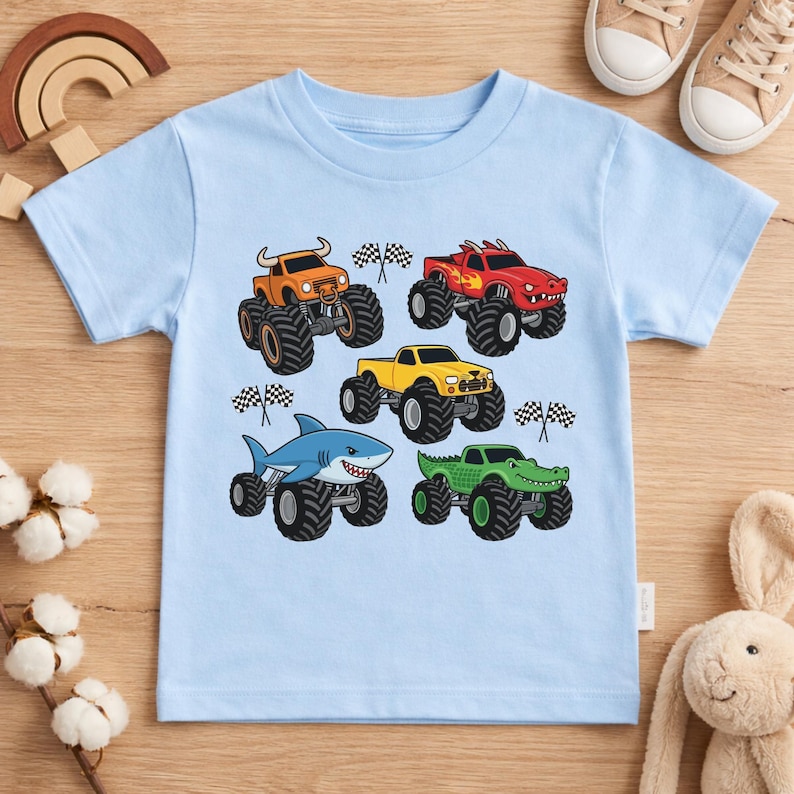 May include: A pale blue t-shirt displaying five monster trucks in orange, red, yellow, blue, and green. Each truck has large black tyres and a unique design, including a shark and an alligator. Checkered flags are also included.