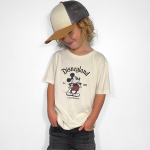 May include: A child wearing a cream-colored t-shirt with a vintage Disneyland graphic featuring Mickey Mouse. The child is also wearing a baseball cap with a brown brim and a gray top.