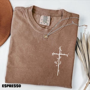 Könnte beinhalten: Espressofarbenes T-Shirt mit weißem floralem Kreuzdesign. Das Shirt hat einen Rundhalsausschnitt und kurze Ärmel. Eine silberne Halskette mit rundem Anhänger liegt auf dem Shirt. Der Text "ESPRESSO" steht unten.