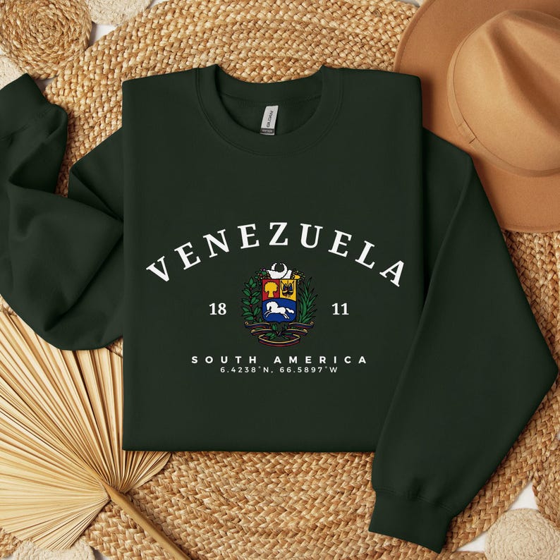 May include: Dark green sweatshirt featuring the word "VENEZUELA" in white arched lettering. Beneath is a colourful emblem, flanked by the numbers "18" and "11". The words "SOUTH AMERICA" and coordinates are below the emblem.