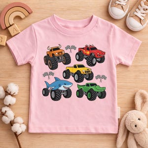 May include: Pale pink short-sleeved t-shirt with five monster truck illustrations. The trucks are orange, red, yellow, a blue shark, and a green alligator. Each truck has large black tyres and a chequered flag.