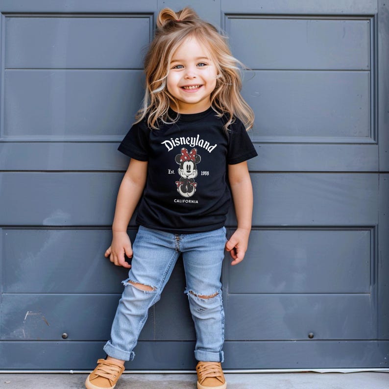 May include: A young child wearing a black t-shirt with the Disneyland logo and a Minnie Mouse graphic. The shirt is paired with light blue ripped jeans and brown shoes. The child is smiling in front of a gray door.