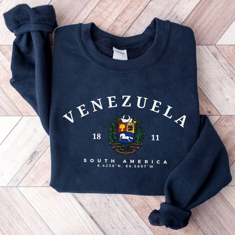 May include: A navy blue sweatshirt featuring the word "VENEZUELA" in white arched lettering. Beneath is a colourful emblem and the text "18 11 SOUTH AMERICA 6.4238&deg; N, 66.5897&deg; W". The sweatshirt rests on a light wood grain surface.