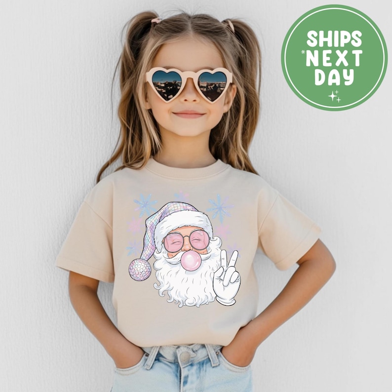 May include: A beige t-shirt with a Santa Claus graphic blowing a bubblegum bubble and giving a peace sign. Santa wears pink sunglasses and a plaid hat. The image also includes the text "SHIPS NEXT DAY" in a green circle.