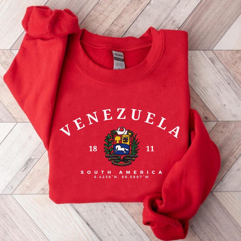May include: A red sweatshirt featuring the word "VENEZUELA" in white. Below is a crest with a coat of arms, the numbers "18" and "11", and the text "SOUTH AMERICA 6.4238&deg; N, 66.5897&deg; W".