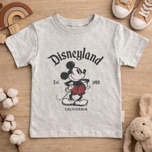 May include: A light gray t-shirt featuring a graphic of Mickey Mouse with the text "Disneyland" above and "Est. 1955 California" below. The cartoon Mickey Mouse is in black, red, and white. The shirt is short-sleeved.