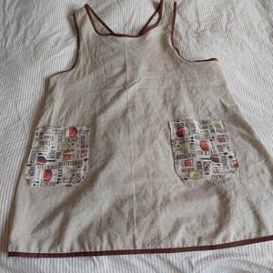 May include: A tan apron with two front pockets featuring a cocktail-themed print. The apron has a brown trim and straps. The pockets display text including "Whiskey", "Cognac", and "Cocktail".