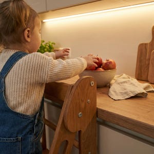 May include: A child in denim overalls and a cream-colored sweater reaches for red apples in a ceramic bowl. A wooden step stool is in the foreground, and wooden cutting boards are in the background.