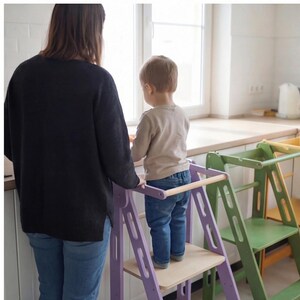 May include: A child stands on a lilac wooden kitchen helper stool with a wooden safety bar. A woman stands nearby. A green and yellow kitchen helper stool is also visible.