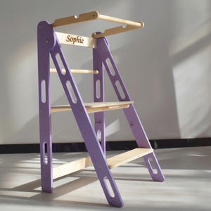 May include: A purple and natural wood learning tower with the name "Sophie" printed on the top. The tower has a ladder design with multiple platforms and a top bar.
