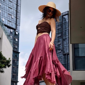 May include: A woman wearing a brown tube top, a long, flowing, ruffled, dusty rose skirt, a straw hat, and sunglasses. The outfit is worn outdoors with tall buildings in the background. The skirt is ankle-length.