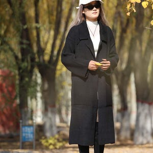 May include: A person wearing a black long coat, white hat, and sunglasses stands outdoors. The coat is knee-length and paired with black boots. The background features trees with yellow leaves, suggesting an autumn setting.