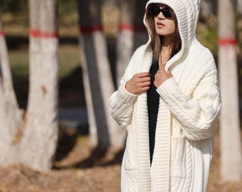 May include: A long, off-white hooded cable-knit cardigan. The sweater has a relaxed fit, long sleeves, and two front pockets. The model is wearing sunglasses and a black top. The background features trees with white trunks and red accents.