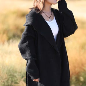 May include: A long, black knitted cardigan with a shawl collar. The cardigan has long sleeves and a relaxed fit. A white top is worn underneath. The background is a blurred field of dry grass.