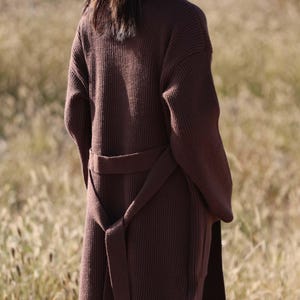 May include: A long, dark brown knit cardigan with a matching belt tied at the waist. The cardigan has long sleeves and a ribbed texture. The model is standing in a field of tall grass.