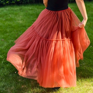 May include: A flowing, tiered, rust-colored tulle skirt. The skirt is paired with a black tank top. The skirt has a gathered waist and a full, airy silhouette, perfect for special occasions or dance. The skirt is shown outdoors on a grassy lawn.