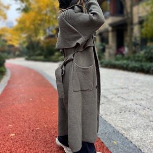 May include: A long, olive green knit coat with a belted waist and large pockets. The coat is worn over dark blue jeans and white sneakers. The person is standing on a red path with trees in the background.