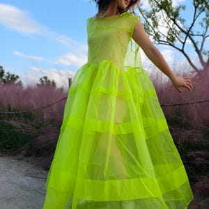 May include: A neon yellow dress with a layered skirt and a sheer overlay. The dress has a round neckline and cap sleeves. The skirt features horizontal bands of the same neon yellow material. The background includes pink foliage and a blue sky.
