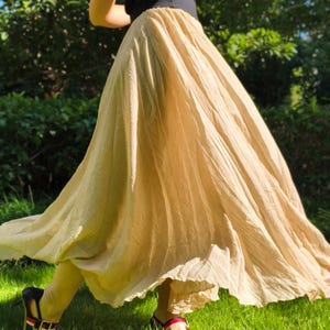 May include: A long, beige skirt with a pleated design. The skirt is worn with a black top and sandals with red, black, and gold details. The image is taken outdoors on a grassy lawn.
