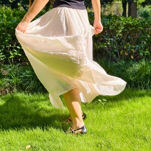 May include: A black sleeveless top paired with a long, flowing, cream-colored skirt featuring a gathered elastic waistband and a textured, lightweight fabric that flares outward, worn with black sandals with thin straps and red accents.