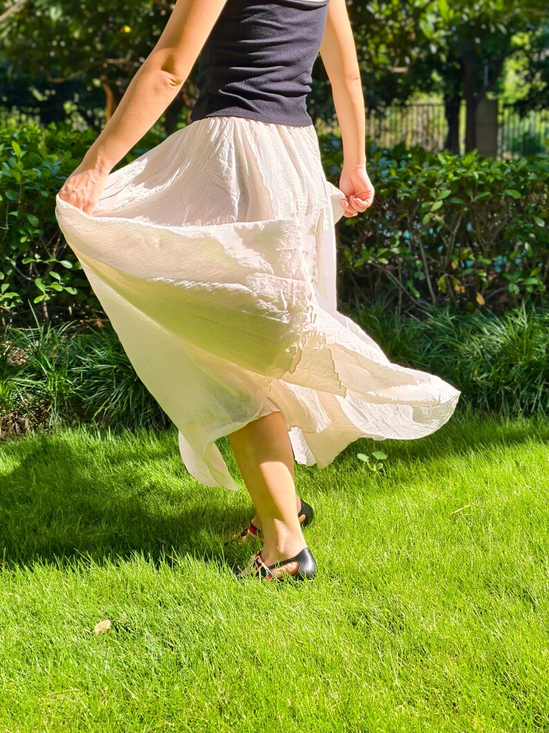 May include: A black sleeveless top paired with a long, flowing, cream-colored skirt featuring a gathered elastic waistband and a textured, lightweight fabric that flares outward, worn with black sandals with thin straps and red accents.