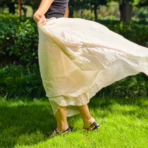 May include: A black sleeveless tank top paired with a flowing, cream-colored midi skirt. The skirt features a lightweight, textured fabric that catches the air, creating a dynamic, flared silhouette that extends outward from the waistline.