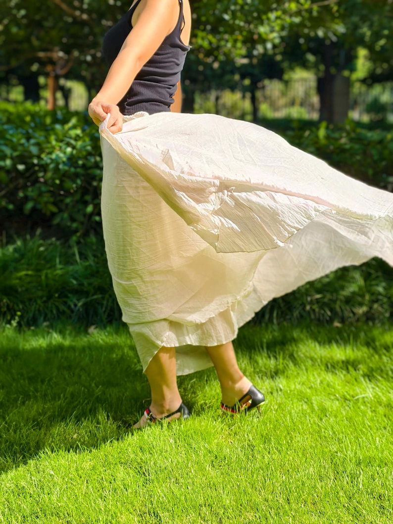 May include: A black sleeveless tank top paired with a flowing, cream-colored midi skirt. The skirt features a lightweight, textured fabric that catches the air, creating a dynamic, flared silhouette that extends outward from the waistline.