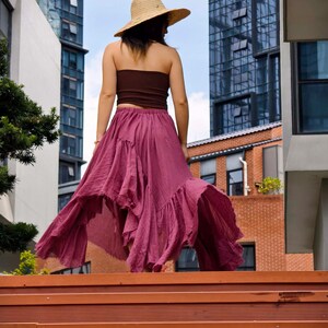 May include: A woman wearing a brown tube top, a flowing magenta skirt, and a straw hat. The skirt has a ruffled hem and is mid-length. The background includes tall buildings and a blue sky.