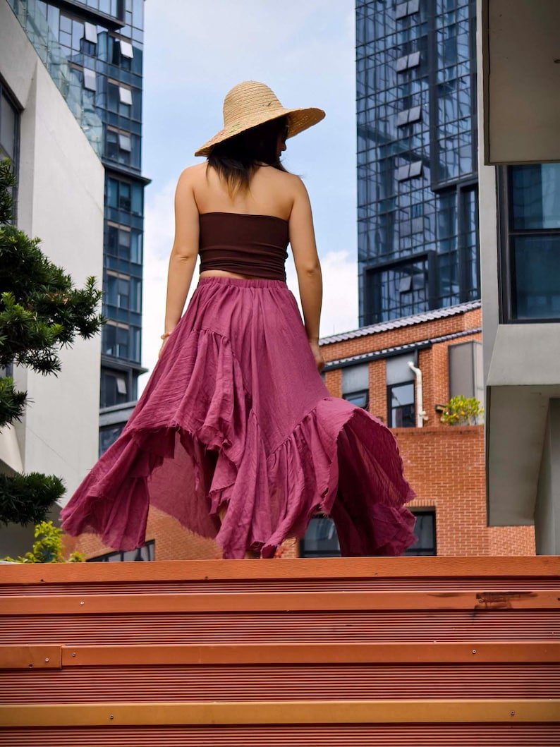 May include: A woman wearing a brown tube top, a flowing magenta skirt, and a straw hat. The skirt has a ruffled hem and is mid-length. The background includes tall buildings and a blue sky.