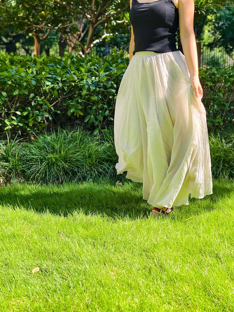 May include: A long, flowing, cream-colored skirt with a crinkled texture and an elasticized waistband, paired with a simple black sleeveless top. The skirt features a full, voluminous silhouette that reaches down to the ankles, creating a soft drape.
