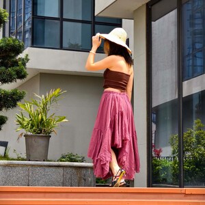 May include: A woman wearing a brown tube top, a flowing, ruffled, dusty rose skirt, and a straw hat. The woman is standing on orange steps, with a building and greenery in the background. She is wearing black sandals with yellow accents.