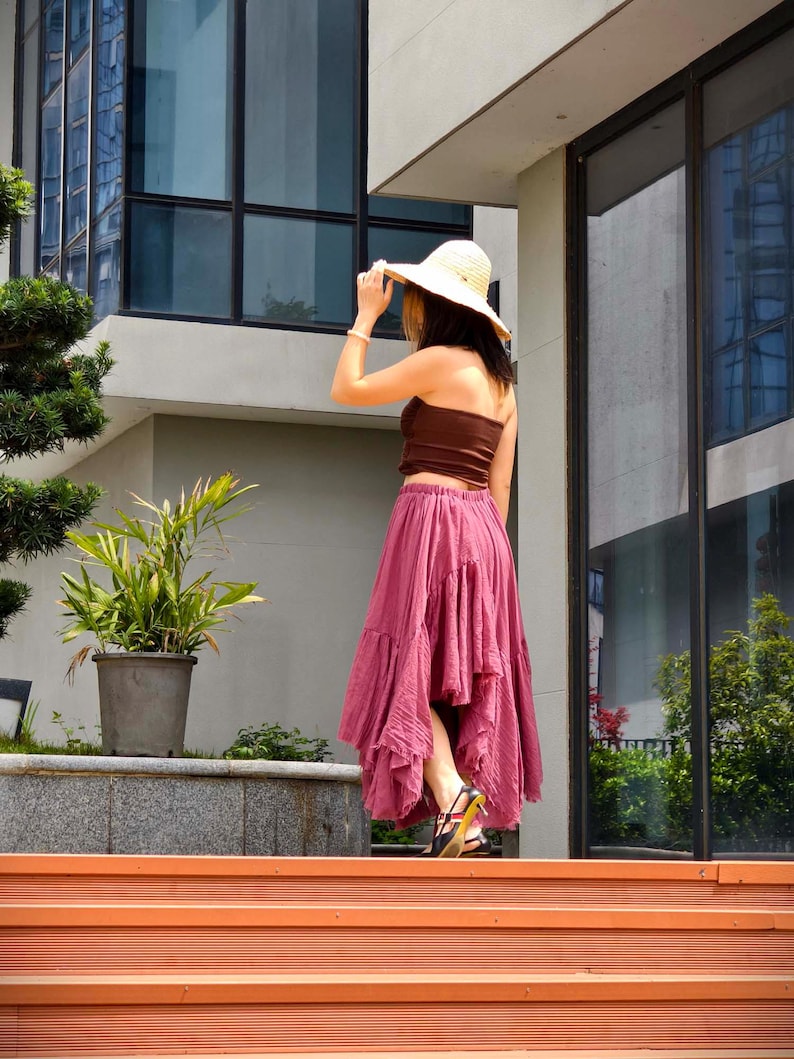May include: A woman wearing a brown tube top, a flowing, ruffled, dusty rose skirt, and a straw hat. The woman is standing on orange steps, with a building and greenery in the background. She is wearing black sandals with yellow accents.