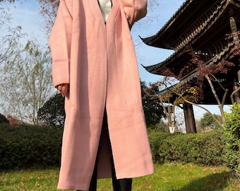 May include: A long, open-front, light pink coat. The coat is made of a soft, textured fabric and has long sleeves. The coat is worn over a white top and black pants. The person is wearing black boots. The background includes a traditional Asian building and green grass.
