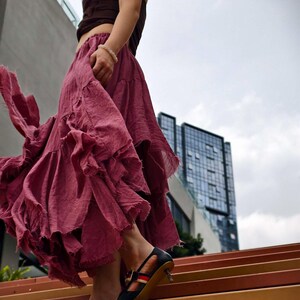 May include: A flowing, ruffled skirt in a deep rose color is paired with a brown top. The outfit is complemented by black heels with red and white stripes. The scene is set outdoors with a modern building in the background.